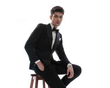 Siyah smokinli yakışıklı damat bir elini cebinde tutarken kollarını kalçasına koyup beyaz arka planda oturuyor.