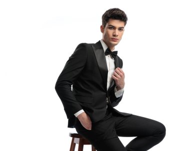 Cebinde siyah smokin olan ve beyaz arka planda oturan seksi, zarif bir adamın portresi.