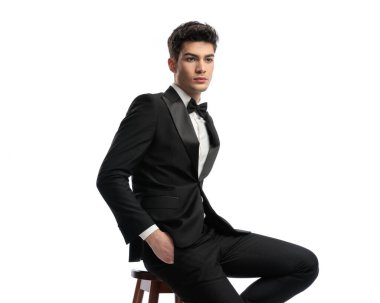 Siyah smokinli, cebinde el ele tutuşan ve beyaz arka planda poz veren zarif bir adamın portresi. 