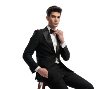 Siyah zarif smokinli, kendine güvenen bir adam, cebinde el ele oturuyor ve beyaz arka planda papyon takıyor.