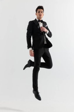 Siyah takım elbiseli moda adamı bacağı bükülmüş ve yakasını gri arka planda düzeltiyor.