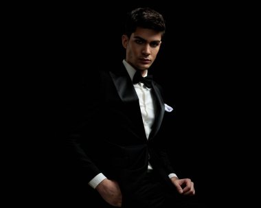 Siyah smokinli ve beyaz gömlekli rahat iş adamı, siyah arka planda cebini tutarken oturuyor.