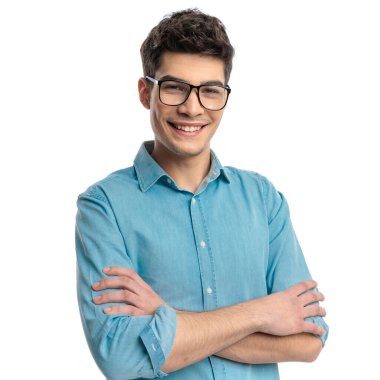 Güneş gözlüklü ve mavi gömlekli genç bir adamın portresi beyaz arka planda kolları bağlı dururken gülümsüyor.