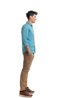 Mavi gömlekli ve kot pantolonlu rahat bir adamın yan görüntüsü beyaz arka planda elleri ceplerinde sırada bekliyor.