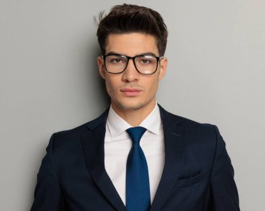 Gözlüklü, mavi kravatlı ve gri arka planda duran siyah takım elbiseli çekici bir moda adamı portresi.
