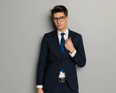 Gözlüklü yakışıklı iş adamının portresi gri arka planda siyah takım elbisesini düzeltiyor.