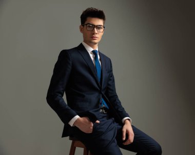 Siyah takım elbiseli genç işadamının yakın plan resmi gri arka planda el ele koltukta dinleniyor.