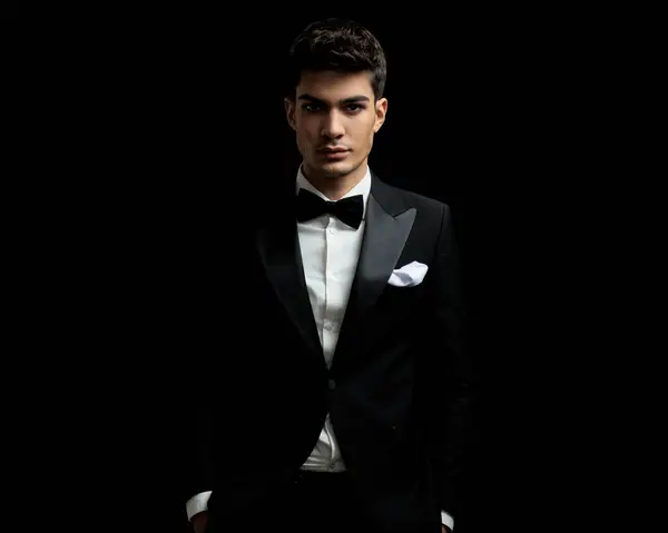 closeup of young man wearing tuxedo and bowtie standing on black background with hands in pockets