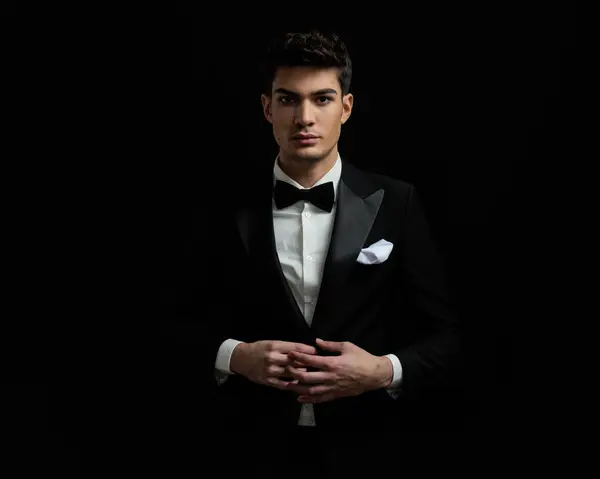 portrait of attractive groom wearing black tuxedo and bowtie standing on black background