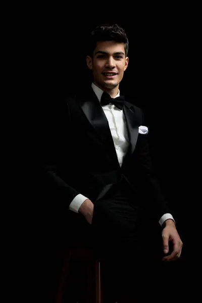 young groom wearing black tux and bowtie is resting on wooden chair while holding pocket on black background