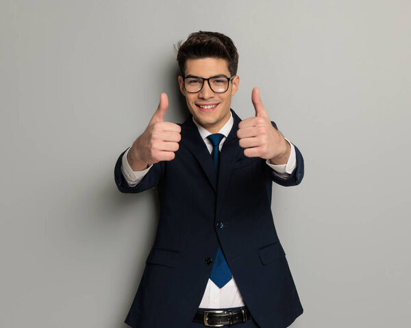 closeup of handsome fashion man making thumbs up sign while standing on grey background in black suit