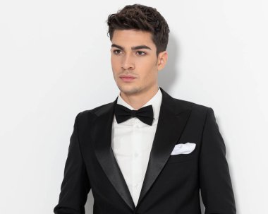 Siyah smokinli yakışıklı damat gözlerini kaçırıp gri arka planda poz veriyor. 