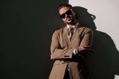 Kahverengi takım elbiseli, güneş gözlüklü yakışıklı bir adam ileriye bakıyor ve gri arka planda kollarını kavuşturuyor.