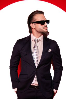 Güneş gözlüklü, cebinde el ele tutuşan ve spot ışıklarının önüne geçmek isteyen zarif, tıraşsız bir adamın portresi. 