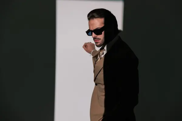 时尚优雅男子的侧视图 戴着太阳镜 触摸墙壁 在灰色背景前摆姿势 图库照片