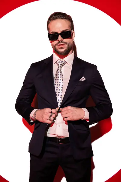 性感优雅男人的肖像 戴着太阳镜调整西服 在聚光灯前摆出一副很酷的姿势 免版税图库图片