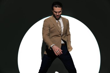 Kahverengi takım elbiseli zarif bir genç, el ele tutuşup spot ışıklarının önünde poz verirken aşağıya bakıyor.