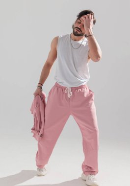 Spor elbiseli, spor ceketli, parmaklarını saçlarında gezdiren ve gri arka planda poz veren kaslı adam.