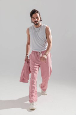 Sakallı, ceketli ve gri arka planda ileri bakan havalı genç kaslı adamın tam vücut resmi. 