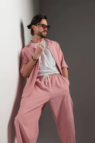 一个留着胡子的性感男人 戴着太阳镜 躺在墙上 边看边摆姿势 手插在灰色背景的口袋里 免版税图库图片