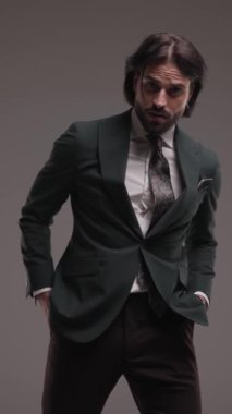 Yakışıklı, uzun saçlı, ceplerinde el ele tutuşan, avuç içi ovuşturan ve gri arka planda rahatça hareket eden zarif bir adam.