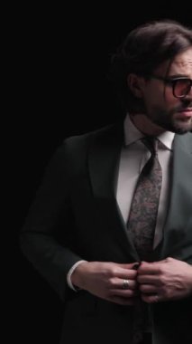 Seksi sakallı iş adamı takım elbisesini ayarlıyor, yan yana bakıyor, el ele tutuşuyor ve siyah arka planda havalı bir şekilde poz veriyor.