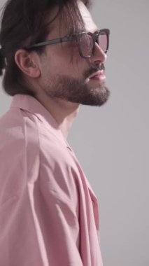 Tıraşsız, pembe gözlüklü ve gri arka planda sakala dokunan seksi bir adamın yan görüntüsü.