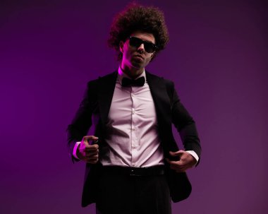 Havalı, kıvırcık saçlı ve güneş gözlüklü moda adamı mor arka planda poz verirken siyah smokini ayarlıyor.