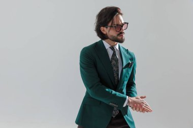 Gri arkaplan önünde avuç içlerine dokunmaya çalışan yeşil takım elbiseli çekici, zarif bir adamın portresi.