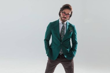 Zarif yeşil takım elbiseli yakışıklı genç adam ceplerinde el ele tutuşmuş, ileriye bakıyor ve gri arka planda poz veriyor.
