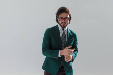 Yeşil takım elbiseli, mutlu zarif bir adamın portresi parmaklarına dokunuyor ve gri arka planda poz veriyor.