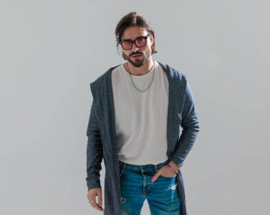 Güneş gözlüklü çekici bir genç adam ceplerinde el ele tutuşuyor ve gri arka planda ileriye bakıyor.