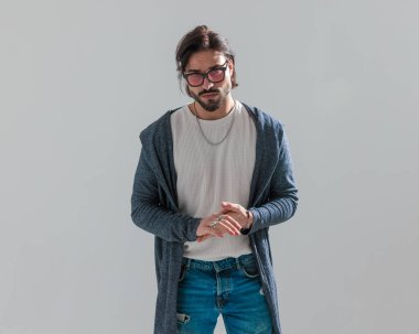 Traş olmamış, güneş gözlüklü, avuçlarını ovuşturan ve gri arka planda ileriye bakan sıradan bir adam. 