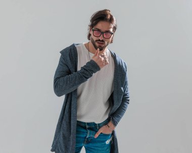 Yakışıklı, sakallı, güneş gözlüklü, el ele tutuşan, çenesine dokunan ve gri arka planda düşünen bir adam.