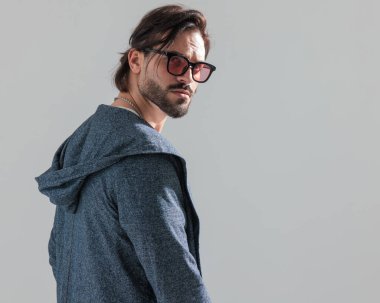 Güneş gözlüklü seksi sakallı adamın arka planı. Arkasına bakıyor ve gri arka planda poz veriyor.