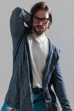 Güneş gözlüklü sakallı genç adam, elini boynunun arkasına koyup gri arka planda poz veriyor. 
