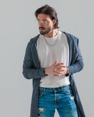 Altın çerçeveli gözlüklü, kendine güveni olmayan, parmaklarına dokunan ve gri arka planda yer almaya çalışan bir adam.