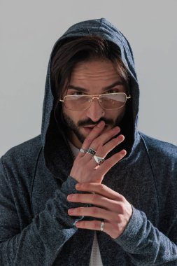 Altın çerçeveli gözlüklü çekici bir adamın portresi parmaklarıyla dudaklara dokunuyor ve gri arka planda ileriye bakıyor.