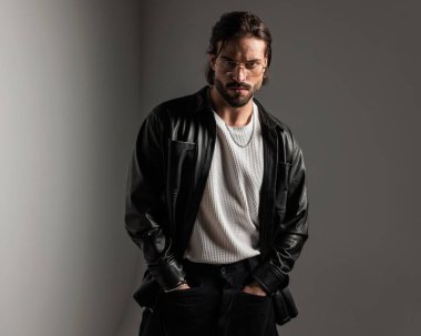 Altın çerçeveli gözlüklü, siyah deri ceketli, gri arka planda el ele tutuşan sert seksi bir adamın portresi.