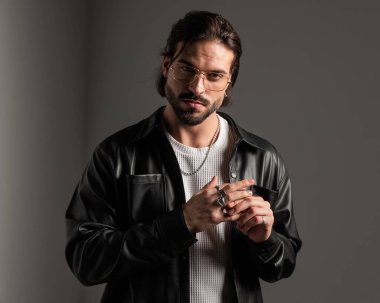 Altın çerçeveli gözlüklü ve deri ceketli havalı moda adamı gri arka planda parmaklarındaki yüzükleri düzeltiyor. 