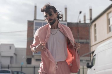 Dikdörtgen güneş gözlüğü takan yakışıklı genç adamın portresi ve şehirde poz verirken pembe günlük gömleği ayarlamak.