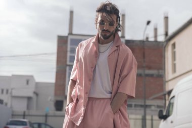 Uzun saçlı, dikdörtgen güneş gözlüklü, pembe günlük elbiseli, el ele tutuşan ve dışarıda poz veren havalı bir adam.