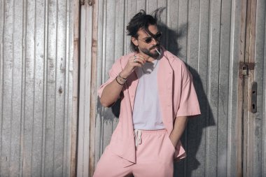 Tıraşsız, pembe ceket ve pantolon giyen, el ele tutuşan, yan yana bakıp sigara içen, dışarıda poz veren seksi bir adam. 