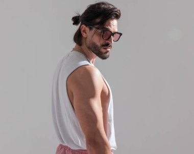 Kendine güveni olan, güneş gözlüklü ve stüdyoda gri arka planda poz veren kaslı bir adamın yan görüntüsü.