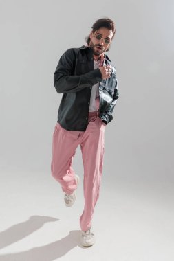 Siyah deri ceketli sakallı, havalı bir adam. Ceplerinde el ele tutuşuyor ve gri arka planların önünde yürüyor.