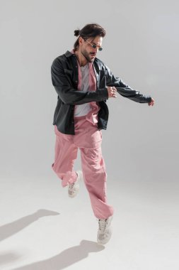 Dikdörtgen güneş gözlüklü modacı havada zıplıyor, yana bakıyor ve gri arka planda poz veriyor.