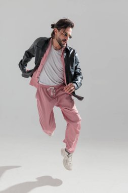 Dikdörtgen güneş gözlüklü, siyah deri ceketli seksi sakallı adam havaya bakıyor, dinamik ve gri arka planda poz veriyor.