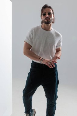 Güneş gözlüklü sıradan bir sakallı adam stüdyoda ayakta durup gri arka planda poz verirken avuçlarını ovalıyor.