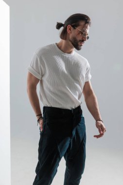 Güneş gözlüklü seksi sakallı adam aşağı bakıyor ve gri arka planda el ele poz veriyor.