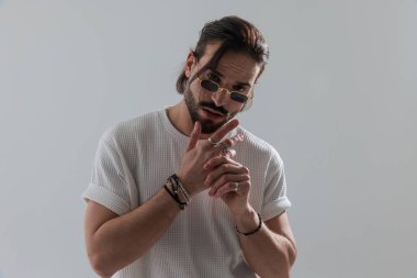 Kendine güveni tam, dikdörtgen güneş gözlüklü, parmaklarına yüzük takan, kameraya bakan ve gri arka planda duran bir adam.